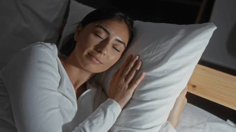 Person in white shirt resting peacefully on bed with white pillow, eyes closed, in minimalist neutral setting